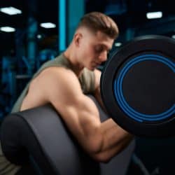 Bodybuilder in sportswear training biceps with barbell on bench, selective focus. Close up of muscular sportsman with perfect body posing in gym in dark atmosphere. Concept of bodybuilding.